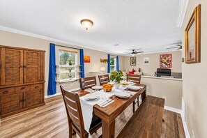 Un coin repas avec une table en bois dressée pour quatre personnes, entourée de chaises, avec un ventilateur de plafond au-dessus et une fenêtre munie de stores.