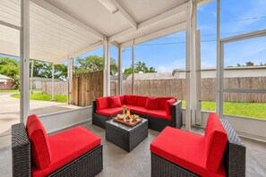 Un patio couvert aménagé avec des meubles en osier et des coussins rouges, doté d’une véranda en verre et offrant une vue sur le jardin arrière.