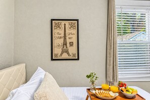 Une chambre d’hôtel avec un lit, un plateau de petit-déjeuner avec des fruits et du jus, un tableau encadré représentant la Tour Eiffel, et une fenêtre avec des stores.