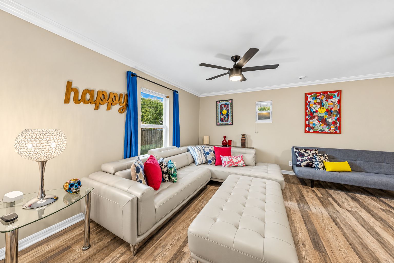 A modern living room with a sectional sofa, a tufted ottoman, a glass coffee table, and a ceiling fan.