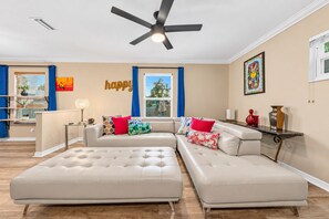 Un salon moderne avec un canapé d’angle, un ventilateur de plafond et une grande fenêtre ornée de rideaux bleus.