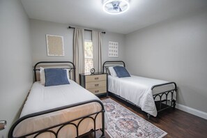 Une chambre avec deux lits simples, une table de chevet, une commode, un tapis à motifs et un ventilateur de plafond.