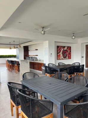 A modern outdoor dining area with a long table and black chairs, a kitchen area with bar stools, and a ceiling fan. - Casa Blue Jade  2 bedrooms loft at Zoho Skies Puerto Vallarta. Roof top pool. (Puerto Vallarta)