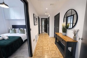 A hotel room with a bed, a bedside table with a vase, a mirror, and a hallway with a geometric patterned floor.