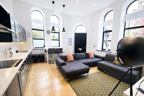 A modern kitchen with a dining area, a sofa set, and large windows.