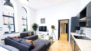 Una sala de estar moderna con un sofá, televisor y plantas. Se observa una cocina con armarios y una encimera.