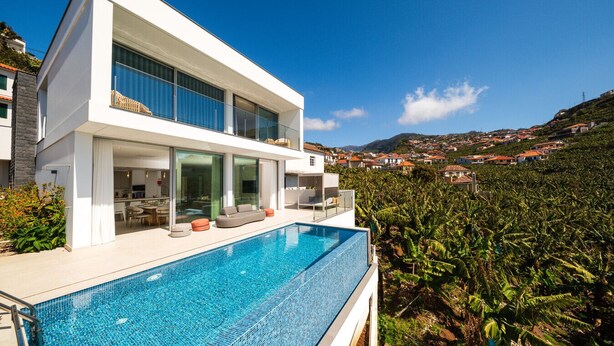 Una casa moderna de dos pisos con piscina, ventanas amplias y vista a un pueblo en la ladera.