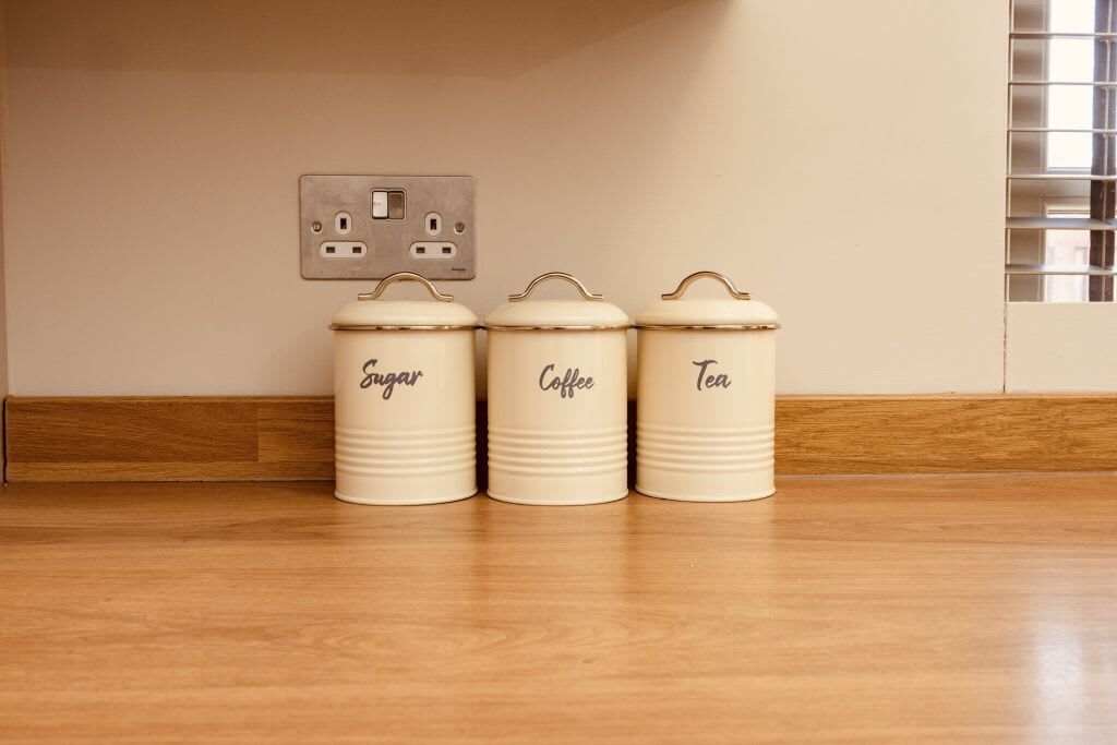 Tres recipientes de cerámica blanca, etiquetados 'Sugar', 'Coffee' y 'Tea', sobre un piso de madera frente a una pared beige con un enchufe.
