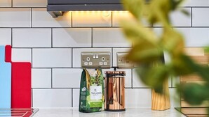 Una cocina con salpicadero de azulejos blancos, una cafetera, una bolsa de café y un objeto rojo y azul.