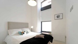 Un dormitorio con cama, lámpara de noche, ventana y un cuadro en la pared.