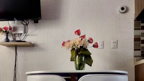 Ein Essbereich mit einem runden Tisch, zwei weißen Stühlen, einer Vase mit Blumen, einem an der Wand befestigten Fernseher und einer Steckdose.