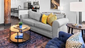 Una sala de estar moderna con un sofá gris, un sillón azul, un tapete estampado y una cocina al fondo.