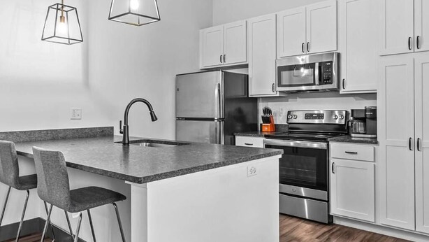 Una cocina moderna con armarios blancos, electrodomésticos de acero inoxidable, encimera de granito y taburetes de bar.