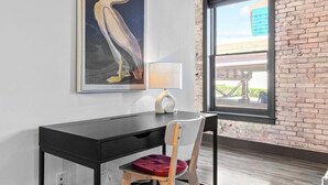 Una habitación con un escritorio, una silla, una lámpara, un cuadro enmarco con un pájaro y un ventanal con vistas a edificios.
