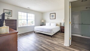 A bedroom with a bed, a dresser, a window, a door leading to a shower, and a framed picture on the wall. - Keystone Garden Villa B (Indianapolis)