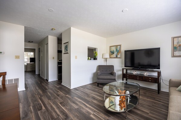 A modern living room with a flat-screen TV, a glass coffee table, and a grey armchair.