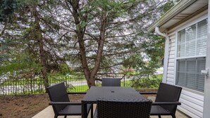 An outdoor patio with a table and chairs, a tree, and a view of a fence and greenery. - Keystone Garden Villa B (Indianapolis)