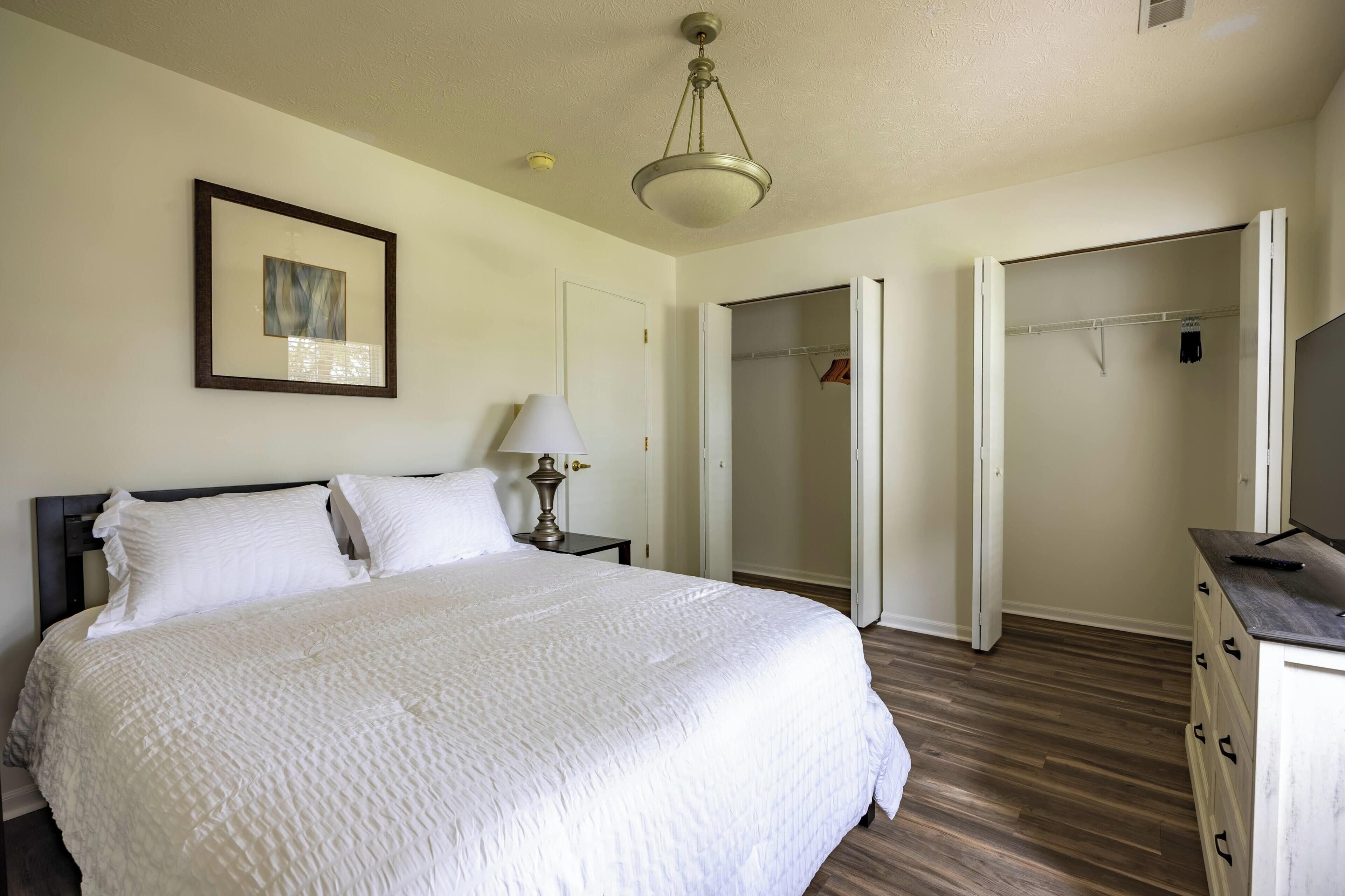A neatly made bed with a white comforter, a bedside lamp, a framed picture on the wall, and a walk-in closet with a mirror.