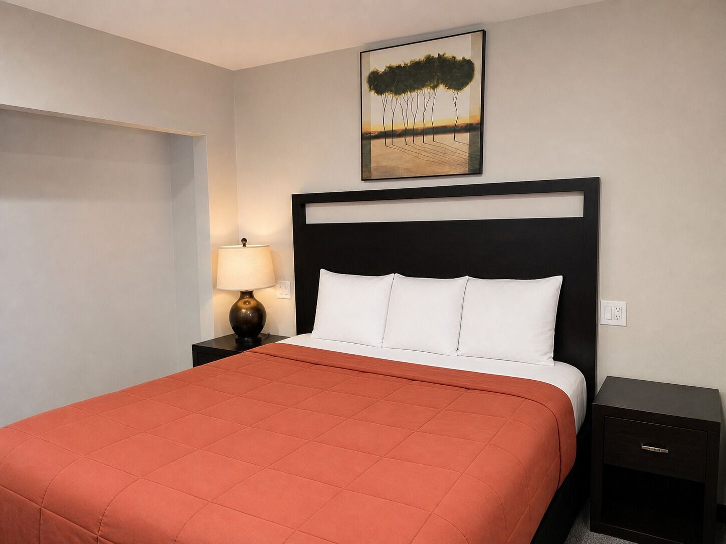 Ein Hotelzimmer mit Bett, Nachttisch mit Lampe, einem Bild an der Wand und einer kleinen Kommode.