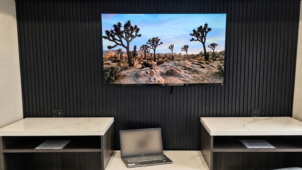 Quarto com duas escrivaninhas de tampo em mármore, um laptop e uma TV fixada em uma parede escura exibindo uma paisagem desértica.