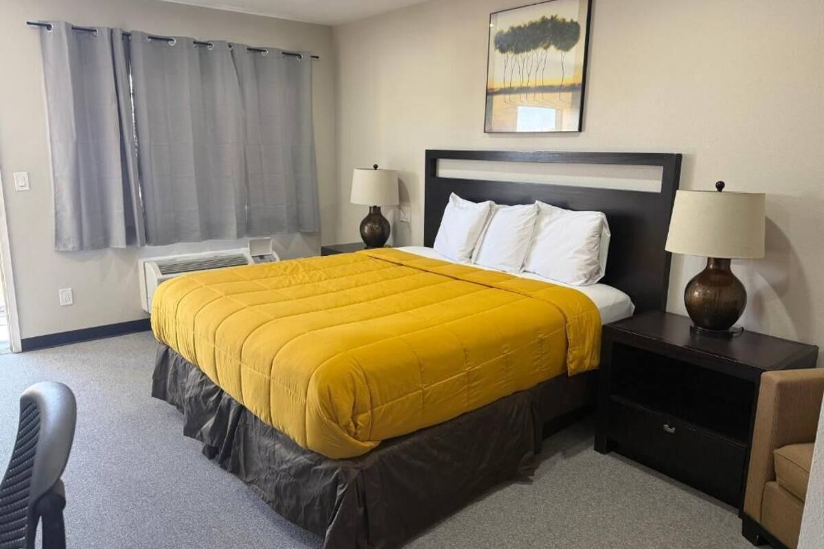 A hotel room with a bed, two bedside lamps, a nightstand, a chair, a window with curtains, and a painting on the wall.