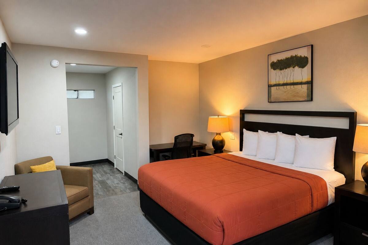 A hotel room with a bed, bedside tables, a desk, a chair, and a painting on the wall.