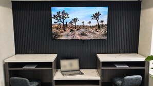 Um espaço de escritório moderno com uma televisão de tela grande fixada em uma parede escura, duas mesas com cadeiras e um laptop em uma delas.