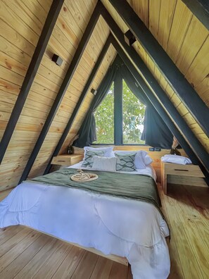 A cozy attic bedroom with a pitched roof, wooden beams, a large window, a bed with pillows, a bedside table, and a nightstand.