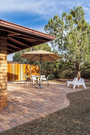 An outdoor patio with a brick floor, a table with chairs, a large umbrella, and white lounge chairs. - Flaming Arrow: Zen Retreat, Close to Hiking/Shops (Sedona)