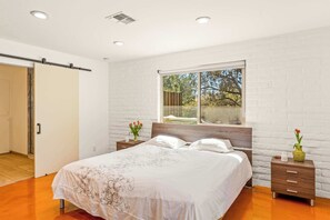 A bedroom with a bed, wooden headboard, white brick walls, a window with a view, a nightstand with a vase, and a sliding door. - Flaming Arrow: Zen Retreat, Close to Hiking/Shops (Sedona)