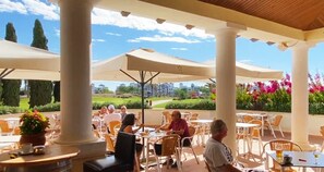 Ein Straßencafé mit Tischen, Stühlen und großen Sonnenschirmen, mit Blick auf einen Golfplatz.