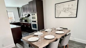 A modern dining area with a wooden table set for four, grey upholstered chairs, and a kitchen in the background. - King William Historic District Cozy 3BR SATX (San Antonio)