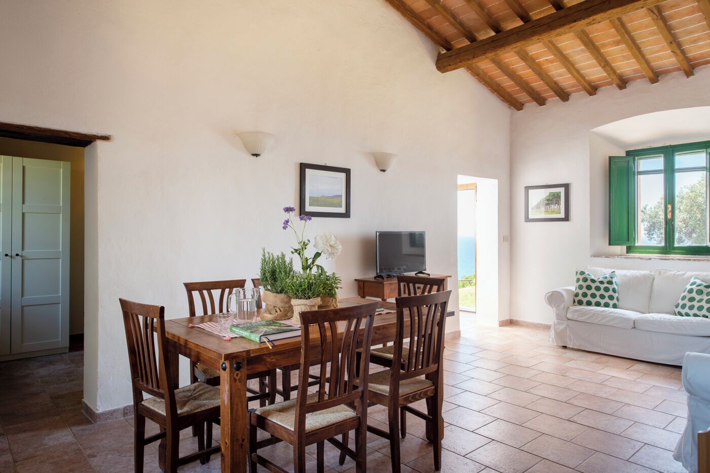 A dining area with a wooden table and chairs, a television, and a view of the sea.