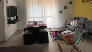 A living room with a TV, a sofa, a dining table with chairs, and a colorful rug.