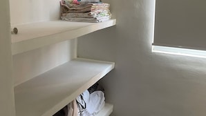 A corner of a room with a white wall, a window, and a metal rod. There are three white shelves, one of which has folded clothes and the other two are empty.