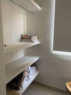 A corner of a room with a white wall, a window, and a metal rod. There are three white shelves, one of which has folded clothes and the other two are empty.