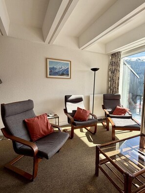 Ein Zimmer mit drei Holzstühlen, einem kleinen Tisch, einer Stehlampe und einem Fenster mit Bergblick.