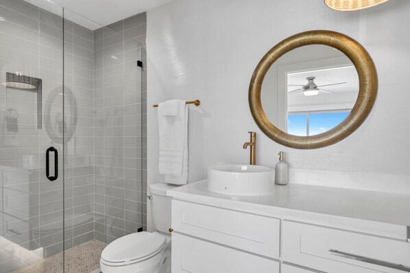 A modern bathroom with a glass shower enclosure, a white toilet, a gold faucet sink, and a large mirror with a gold frame.