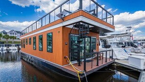 Ein schwimmendes Hausboot mit einer Glasoberdeck und einem Holzboden, sowie ein vor Anker liegendes Boot im Hintergrund.