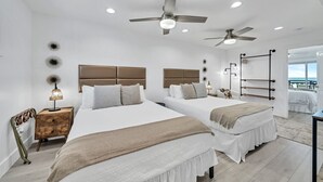 Hotelkamer met twee bedden, plafondventilatoren, een nachtkastje met lamp, een houten nachtkastje en uitzicht op de oceaan.