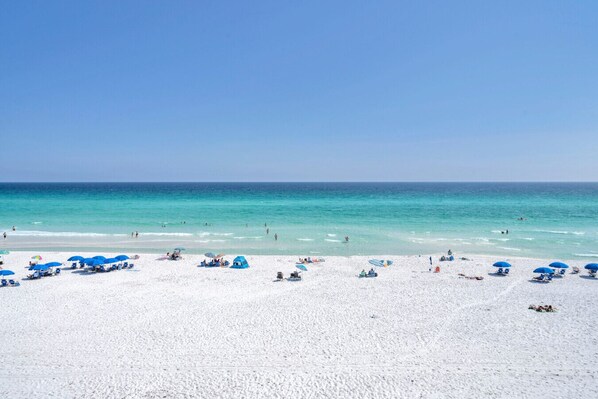 Une plage de sable blanc, avec une eau d’un bleu limpide et des parasols bleus.