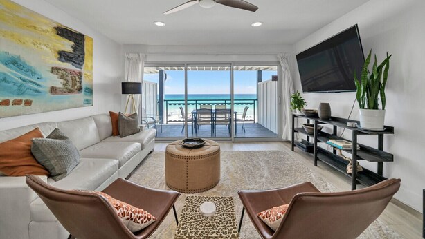 Een woonkamer met een bank, twee stoelen, een salontafel, een plafondventilator, een televisie, en een balkon met uitzicht op de oceaan.