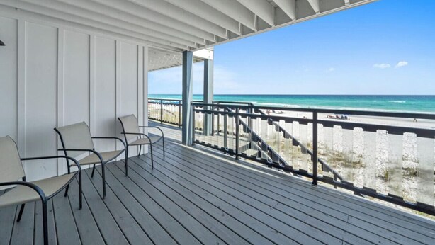 A covered outdoor area with a view of the beach, featuring a wooden deck, chairs, and a railing. - Beachfront | Top-Rated | Sleeps 10 | Seabatical #3 (Destin)
