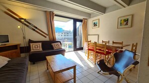 Ein Zimmer mit einer Couch, einem Holztisch, einem Esstisch mit Stühlen, einem Fernseher und einem Balkon mit Blick auf Gebäude.