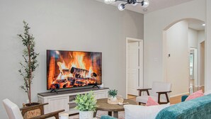 Una sala de estar con televisor mostrando una chimenea, una mesa de comedor con sillas, un sofá y una planta en maceta.