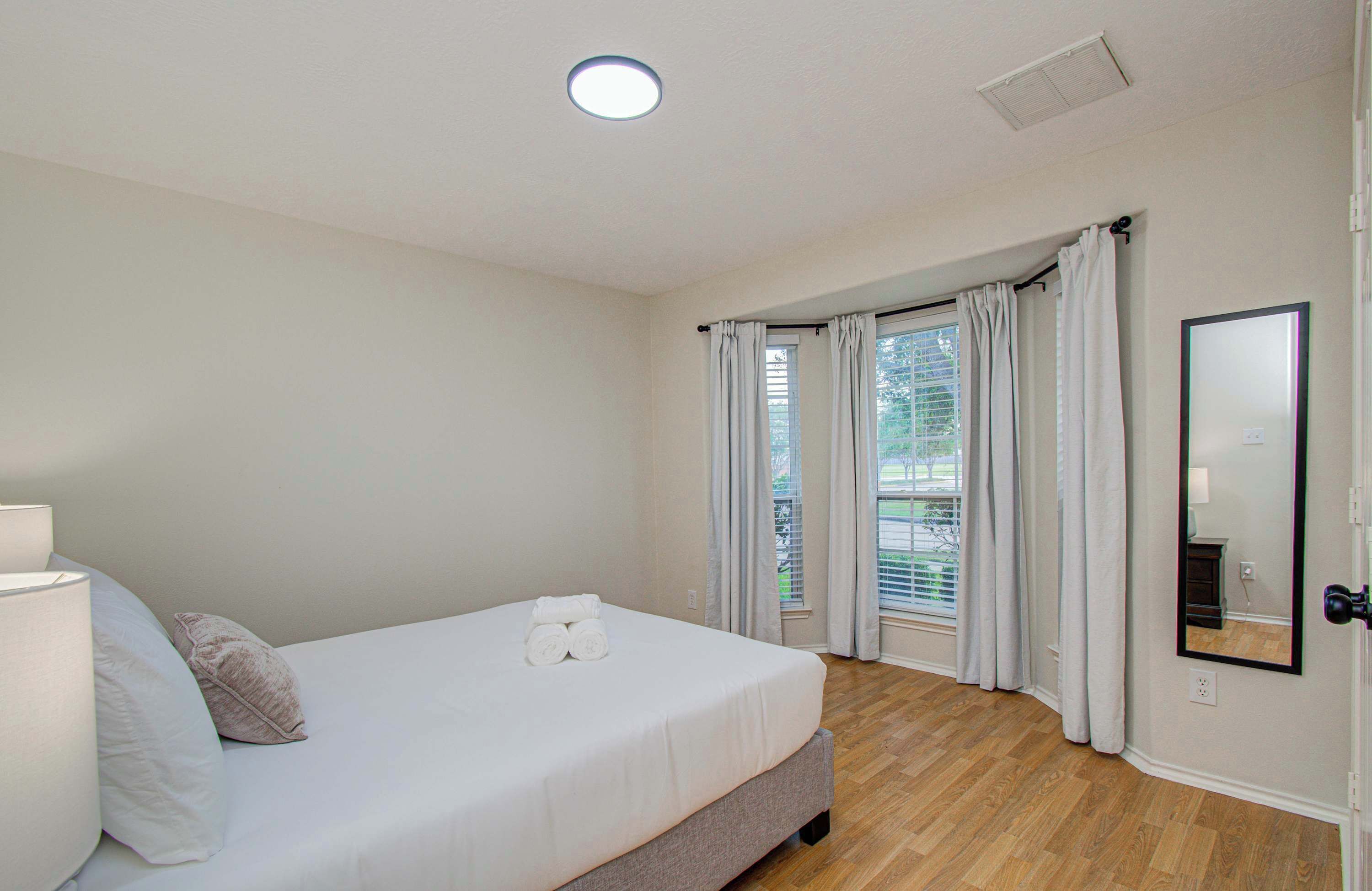 Ein Schlafzimmer mit Bett, Nachttischlampe, einem Fenster mit Vorhängen, einem Spiegel und einer Tür.