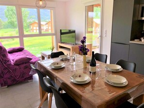 Ein Essbereich mit einem Holztisch, gedeckt für sechs Personen, einem Fernseher und Blick auf eine grüne Landschaft durch große Fenster.