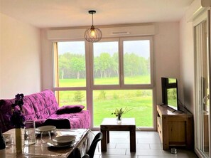 Ein Zimmer mit einem violetten Sofa, einem Esstisch und einem Fernseher auf einem Holzgestell.