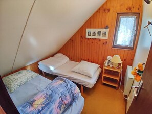 Ein kleiner, gemütlicher Raum mit zwei Betten, einer Holzbalkendecke, einem Fenster mit Spitzenvorhängen und einem kleinen Tisch mit Lampe und Stofftier.