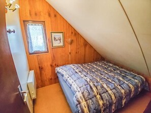 Ein kleines, gemütliches Schlafzimmer im Dachgeschoss mit schrägen Wänden, einem Einzelbett, einem Fenster mit Vorhang und einem gerahmten Bild an der Wand.